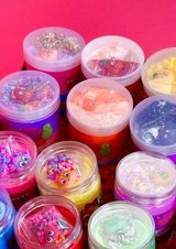Close up of the tops of jars of putty with different coloured charms and sprinkles on a pink background