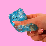 A blue sugar filled fidget toy in the shape of a bear being squashed in a hand, against a pink background