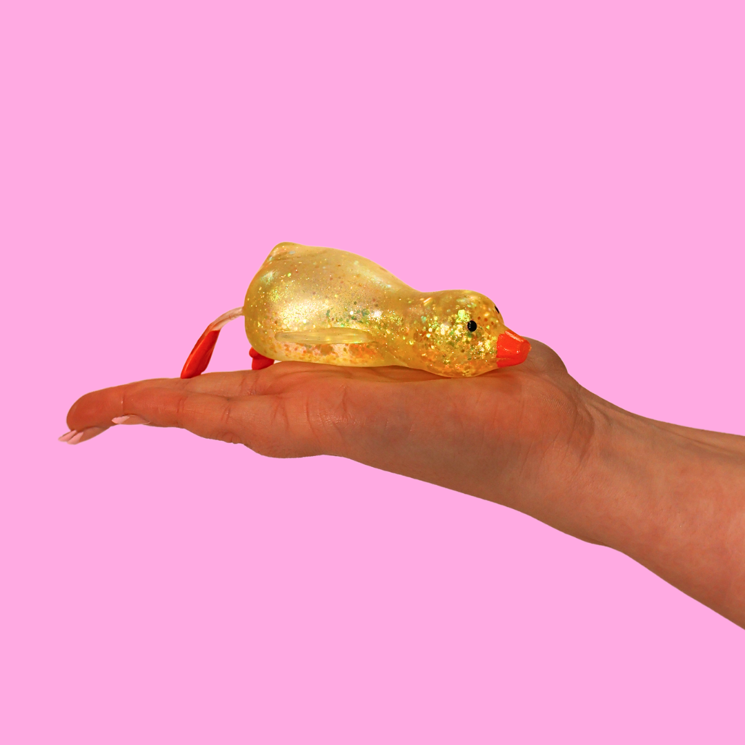 A yellow sugar filled fidget toy, in the shape of a duck, laid on the palm of a hand against a pink background