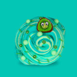 A teal and light green swirled sensory putty decorated with small kiwi slices, green clay sprinkles and green floam balls. A cartoon kiwi charm is on the top with big eyes and a smile