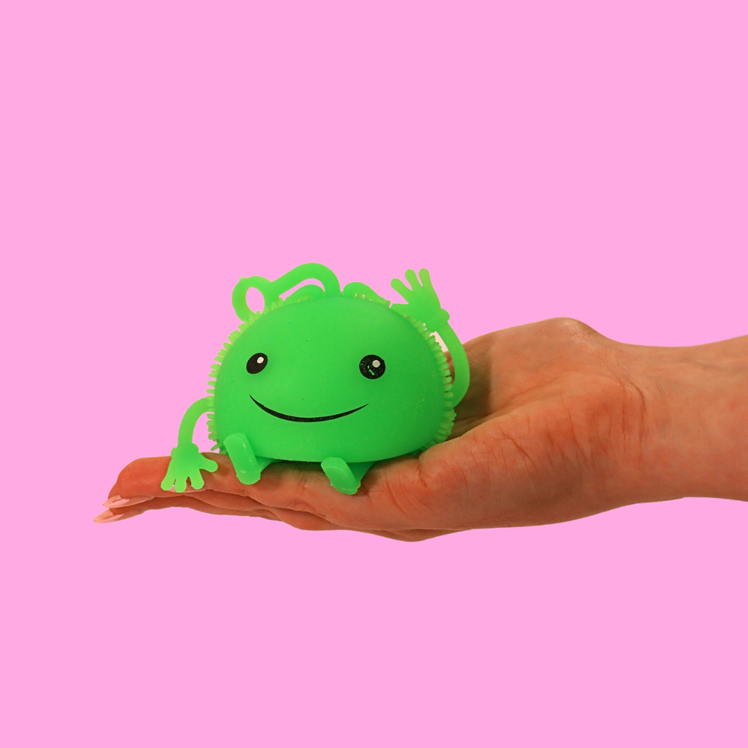 A hand holding a green light up fidget toy that has a smiley face on it, against a pink background