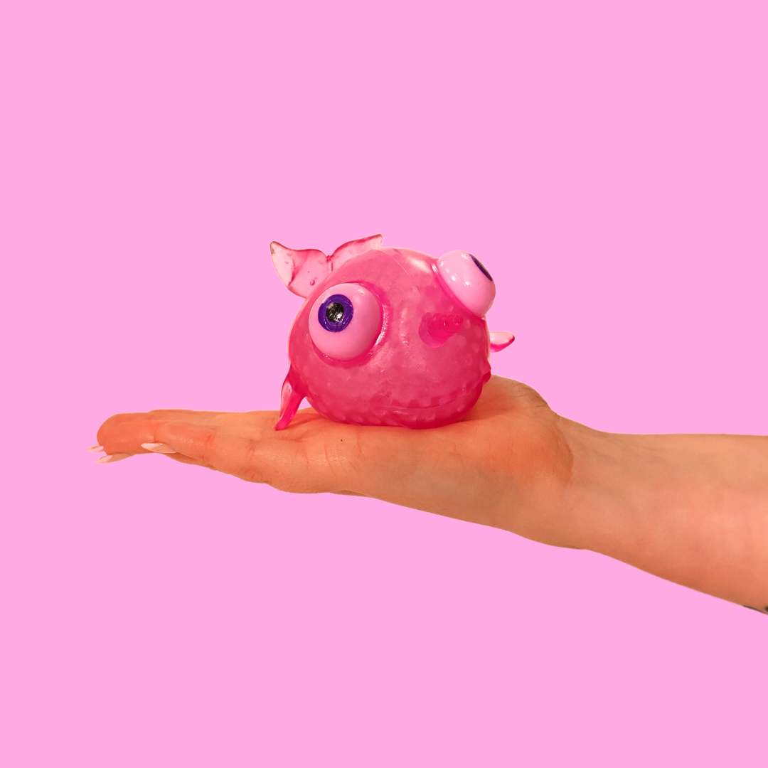 A pink big eyed narwhal fidget toy, in the palm of a hand against a pink background