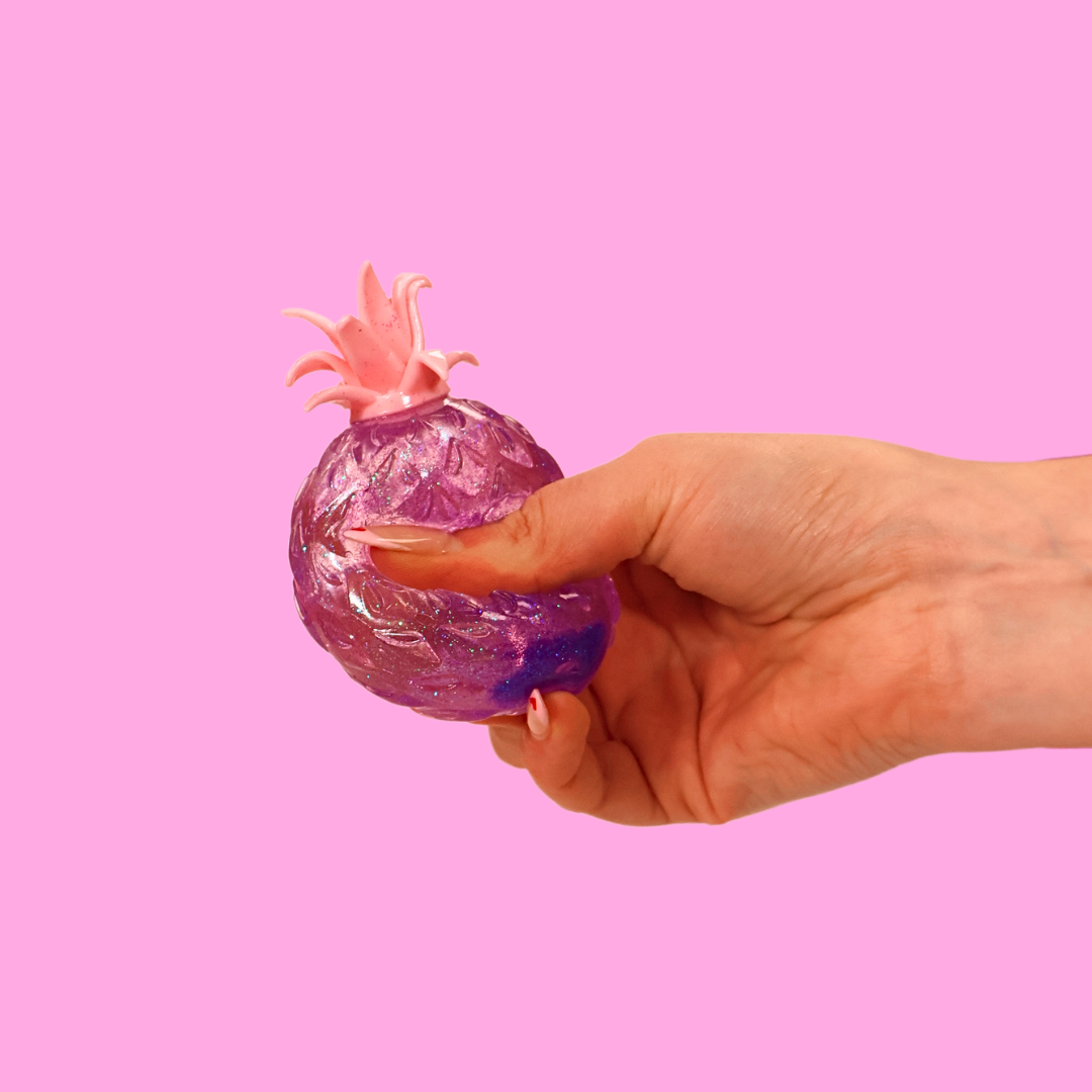 A purple sugar filled fidget toy being squished in a hand, against a pink background