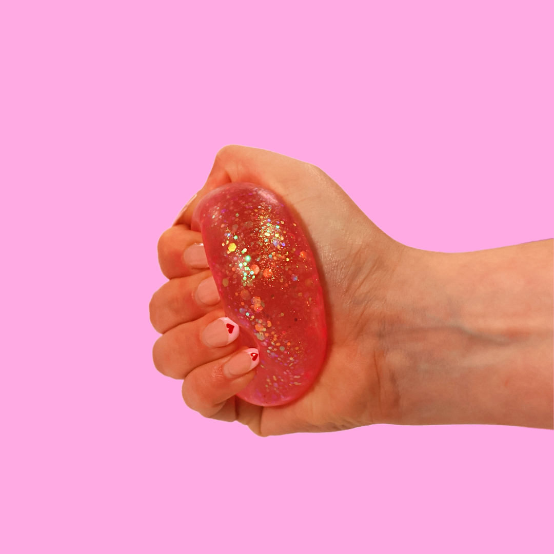 A pink sugar filled fidget toy, being squashed by a hand against a pink background