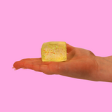 A hand holding a yellow sugar fidget toy in the shape of a block, on a pink background.