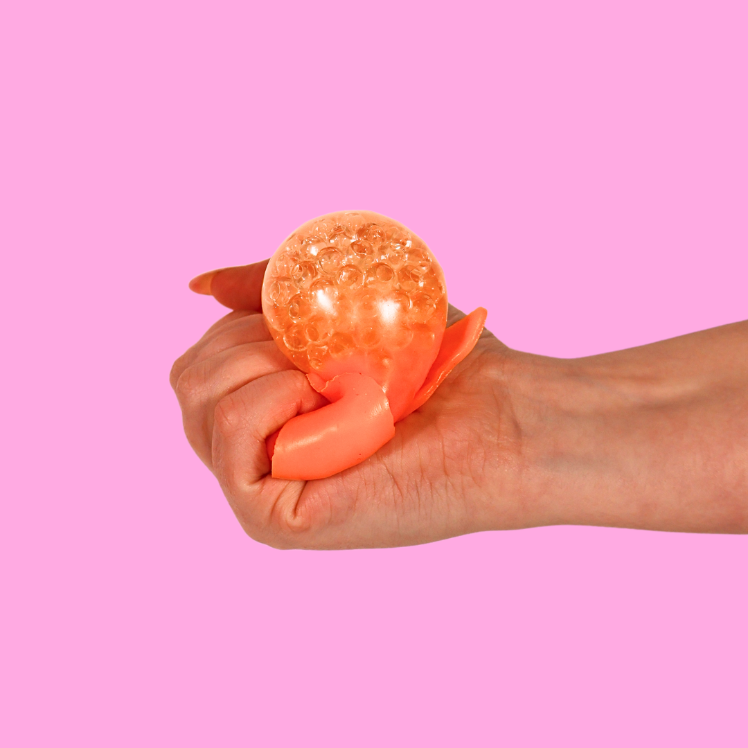 An orange orbs filled fidget toy in the shape of a tangerine being squished in a hand on a pink background 